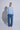 A woman stands with her back to the camera wearing a light blue long-sleeve top and wide-leg blue jeans against a light gray studio background with a prominent shadow.