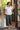 Woman in white top and green pants standing in a doorway with plants outside.