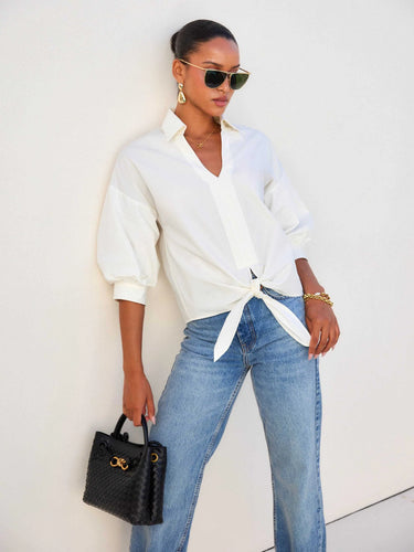 A woman modeling a cream-colored, oversized button-down shirt tied at the waist, paired with blue denim jeans, set against a plain white studio background.