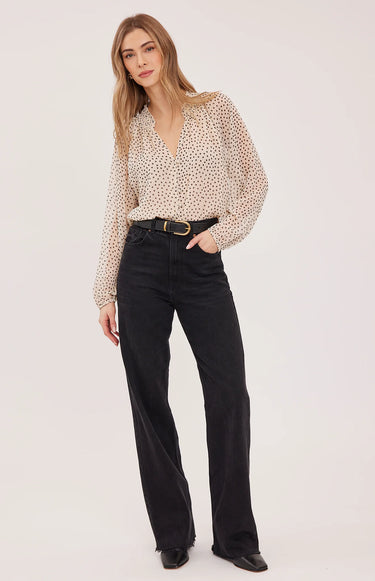 A woman wearing a black polka dot and cream blouse with black jeans on a white background.