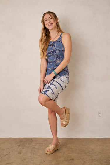 Woman wearing a blue tie-dye dress standing against a plain wall.