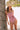 A woman leaning on a tree, modeling a red and white geometric patterned strapless one-piece swimsuit with a matching tied belt, with a sandy beach background