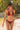 Woman wearing a merlot halter bikini with an O-ring detail and tie-side bottoms, standing on a sandy beach next to clear water and lush green trees, holding an apple.