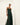 Full-length image of a woman wearing a dark green strapless maxi dress with subtle vertical stripes, a central bust cutout with a knotted detail, and high side slits. She is posing confidently against a white background, wearing brown sandals and wooden bracelets.