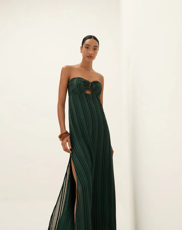 Full-length image of a woman wearing a dark green strapless maxi dress with subtle vertical stripes, a central bust cutout with a knotted detail, and high side slits. She is posing confidently against a white background, wearing brown sandals and wooden bracelets.