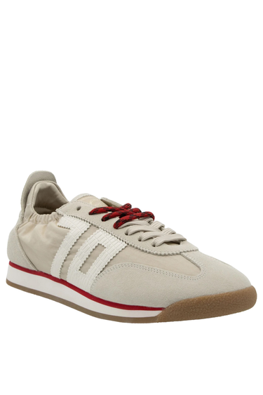 A single beige retro sneaker with white side stripes, a red midsole accent, and red-speckled laces, featuring a suede and nylon upper with a gum rubber sole, presented on a black background.