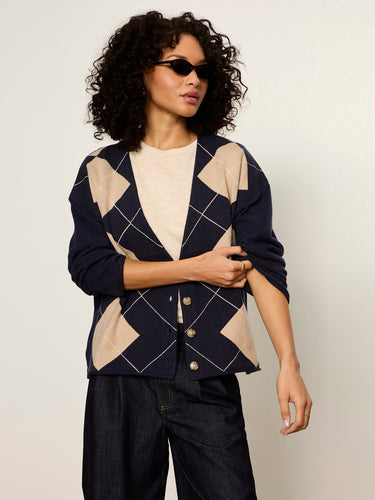 A woman modeling a navy blue and beige argyle cardigan with white geometric lines, worn over a cream t-shirt and dark jeans against a light gray studio background.