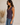 A woman wearing a sleeveless, deep purple ruched V-neck top and blue jeans, standing in a studio setting with a neutral beige background and a decorative vase of pampas grass on the left.