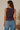 A woman shown from the back wearing a sleeveless dark purple tank top and light blue jeans, standing in a softly lit indoor setting with a neutral wall and a small plant in a vase.