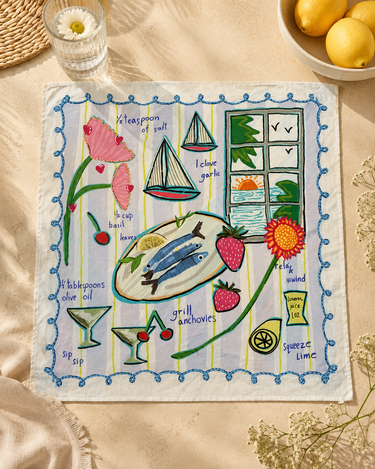 A square embroidered bandana with a colorful Mediterranean-themed design, laid flat on a beige fabric surface with styled props including lemons, a glass, and dried flowers.