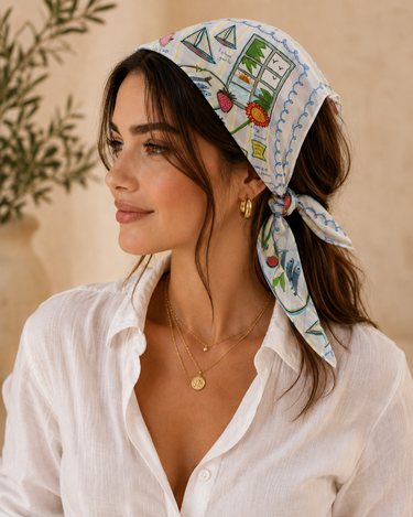 A woman wearing a white cotton headscarf with a colorful beach-themed print, styled with gold jewelry and a white button-down shirt against a warm beige background.