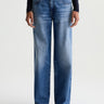 A person wearing a pair of medium-wash blue denim jeans with a straight-leg fit, paired with a black long-sleeve top and dark brown shoes, shown against a plain light gray studio background.