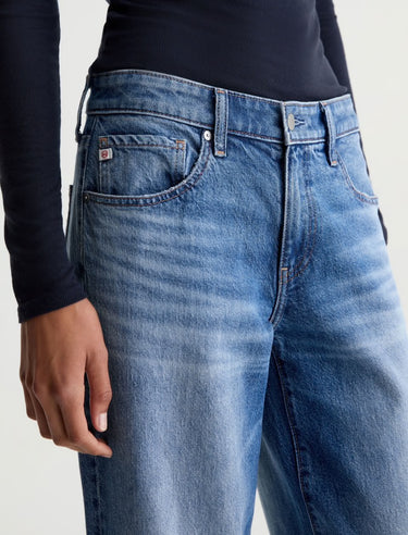 Close-up of a person wearing blue denim jeans with a faded wash and a black long-sleeve top, shown against a light neutral background.