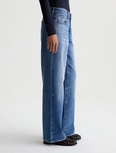 A person shown from the waist down wearing wide-leg blue jeans, a black long-sleeve top, and black shoes against a clean light gray studio background.