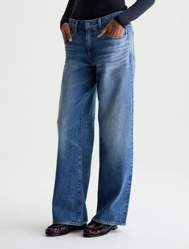A person wearing blue wide-leg denim jeans with a dark navy long-sleeve top and dark brown shoes, standing against a plain light gray studio background.