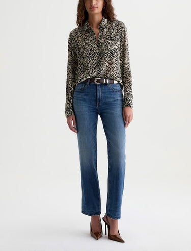Woman wearing a patterned blouse and mid-rise medium blue wash straight leg denim with a distressed hem, fading, and gold stitching on a white background.