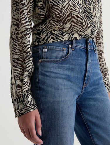 A woman wearing mid-rise medium blue wash straight leg denim with a distressed hem, fading, and gold stitching on a white background.