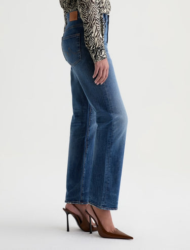 A woman wearing mid-rise medium blue wash straight leg denim with a distressed hem, fading, and gold stitching on a white background.