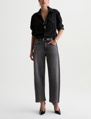 A woman  wearing  dimensional ashy black wash jeans, with contrasting paneled detailing and a black top with black heels on a white background.