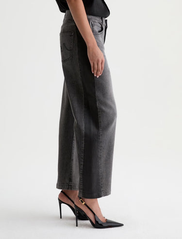 A woman facing the side wearing  dimensional ashy black wash jeans, with contrasting paneled detailing and a black top with black heels on a white background.