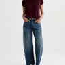 A woman wearing high rise barrel jeans in medium wash with a short sleeve maroon tee and black loafers on a white background.