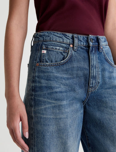 A woman wearing high rise barrel jeans in medium wash with a short sleeve maroon tee on a white background.