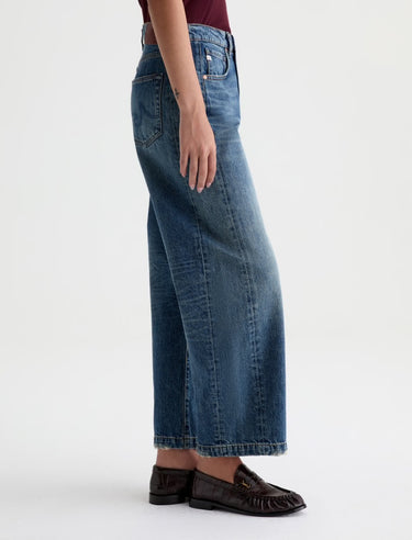 A woman facing sideways in high rise barrel jeans in medium wash with a short sleeve maroon tee and black loafers on a white background.