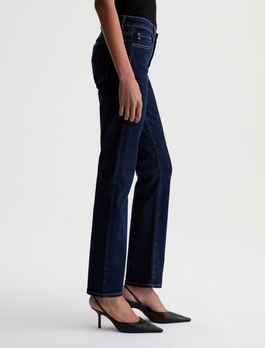 A woman facing sideways wearing low rise straight dark blue jeans and black heels on a white background.