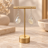 A pair of teardrop-shaped dangle earrings with iridescent shell-like drops and silver wire hoops, displayed on a gold jewelry stand on a beige stone surface.