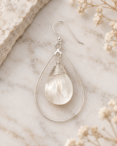 A silver teardrop dangle earring featuring a clear, feathered quartz crystal, styled on a light marble surface with dried flowers and a stone slab.