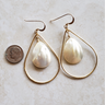 A pair of gold teardrop hoop earrings with white mother-of-pearl teardrop pendants, displayed on a light beige textured surface next to a US dime for scale.