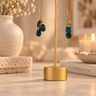 Gold-tone hoop earrings with stacked teal blue gemstone drops, displayed on a gold stand on a marble surface with a warm, softly blurred home decor background.