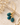 A pair of gold hoop earrings with faceted teal-blue stone drops, styled on a textured beige stone slab with dried white flowers.