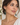 A close-up of a woman wearing gold drop earrings with dark teal beads, shown from the chest up against a softly blurred indoor background.