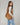 A woman wearing a light beige long-sleeve top and light blue jeans carries a large brown suede shoulder bag against a plain light gray studio background.