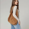 A woman wearing a light beige long-sleeve top and light blue jeans carries a large brown suede shoulder bag against a plain light gray studio background.