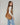 A woman wearing a light beige long-sleeve top and light blue jeans carries a large brown suede shoulder bag against a plain light gray studio background.