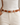 A close-up of a person wearing a slim brown leather belt with a polished silver-tone buckle, paired with off-white jeans and a white ribbed top.