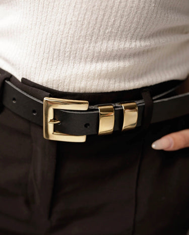 A close-up of a black leather belt with a polished gold-tone buckle, worn with dark trousers and a white ribbed top.
