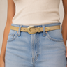 A close-up of a model wearing a light beige leather belt with a polished gold-tone buckle, styled with light-wash blue jeans and a cream ribbed tank top.
