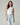 A woman modeling a sleeveless white mock neck top paired with light-wash blue jeans and a white belt, standing against a solid light gray studio background.