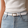 A close-up of a model wearing a slim white leather belt with a gold-tone rectangular buckle, styled with light-wash blue jeans and a white ribbed top.