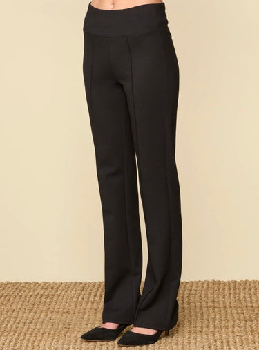 Black high-waisted straight-leg pants shown on a model from the waist down, paired with black pointed-toe heels on a textured beige woven rug against a solid beige background.
