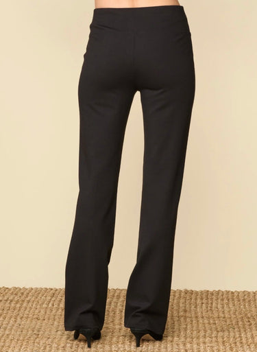 Black wide-leg flare pants shown from the back on a model, paired with black high heels, set against a beige wall and a textured tan rug.