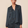 A woman modeling a dark green satin long-sleeve top with a drawstring neckline, paired with matching satin pants, against a solid beige background.