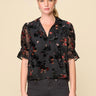 A woman wearing a velvet floral mesh blouse and black jeans on a neutral background.