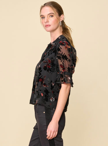 A woman wearing a velvet floral mesh blouse and black jeans on a neutral background.