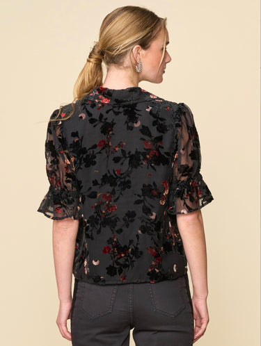 A woman wearing a velvet floral mesh blouse and black jeans on a neutral background.