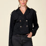 A woman modeling a black double-breasted cropped jacket with silver buttons, paired with dark black jeans, against a solid light beige background.