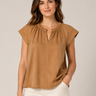 A woman wearing a tan short-sleeve blouse with a split neckline and pleated details, paired with off-white pants, against a neutral beige background.
